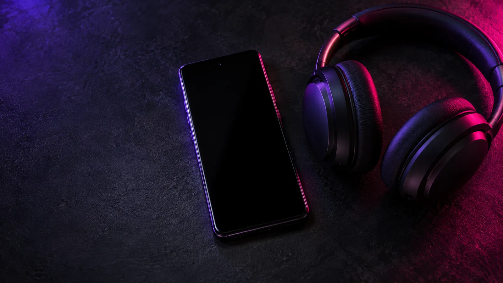 Auriculares junto a un teléfono con la pantalla apagada, sobre una mesa con luz violeta.